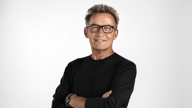 Uomo anziano con braccia conserte e occhiali, sorridente.