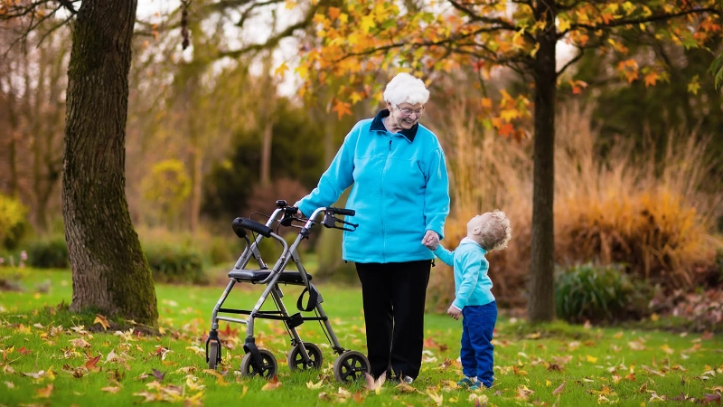 Dans un jardin automnal, une grand-mère en déambulateur tient la main de son petit-fils et lui sourit.