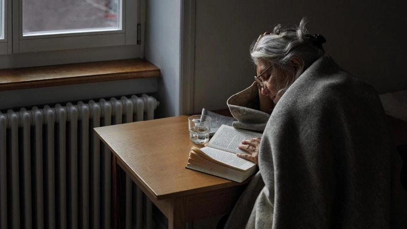 Un anziano con una coperta e un tè legge un libro.