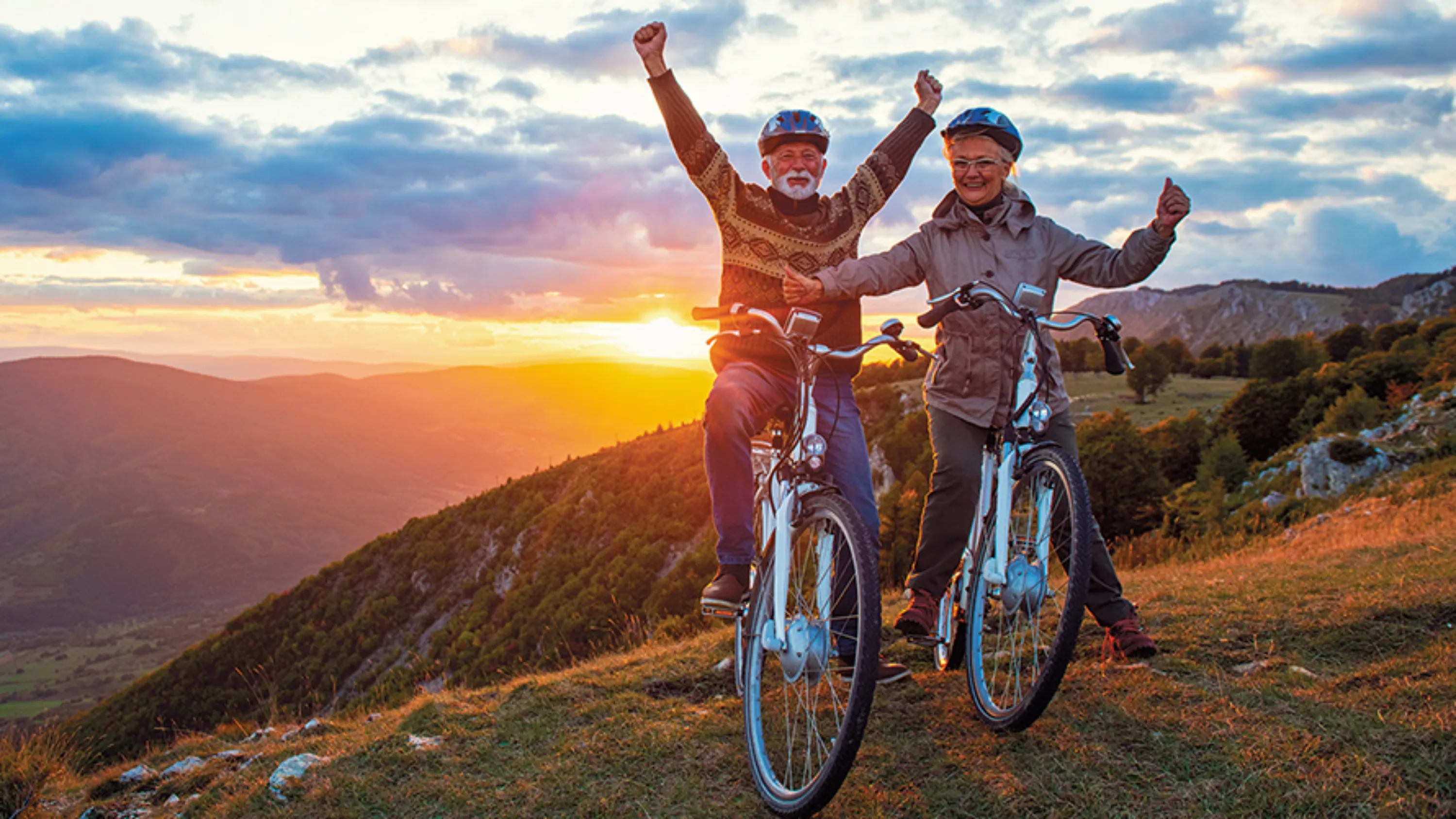 Senioren auf dem Velo stehend, Hände in die Höhe, Freude mit Sonnenuntergang im Hintergrund