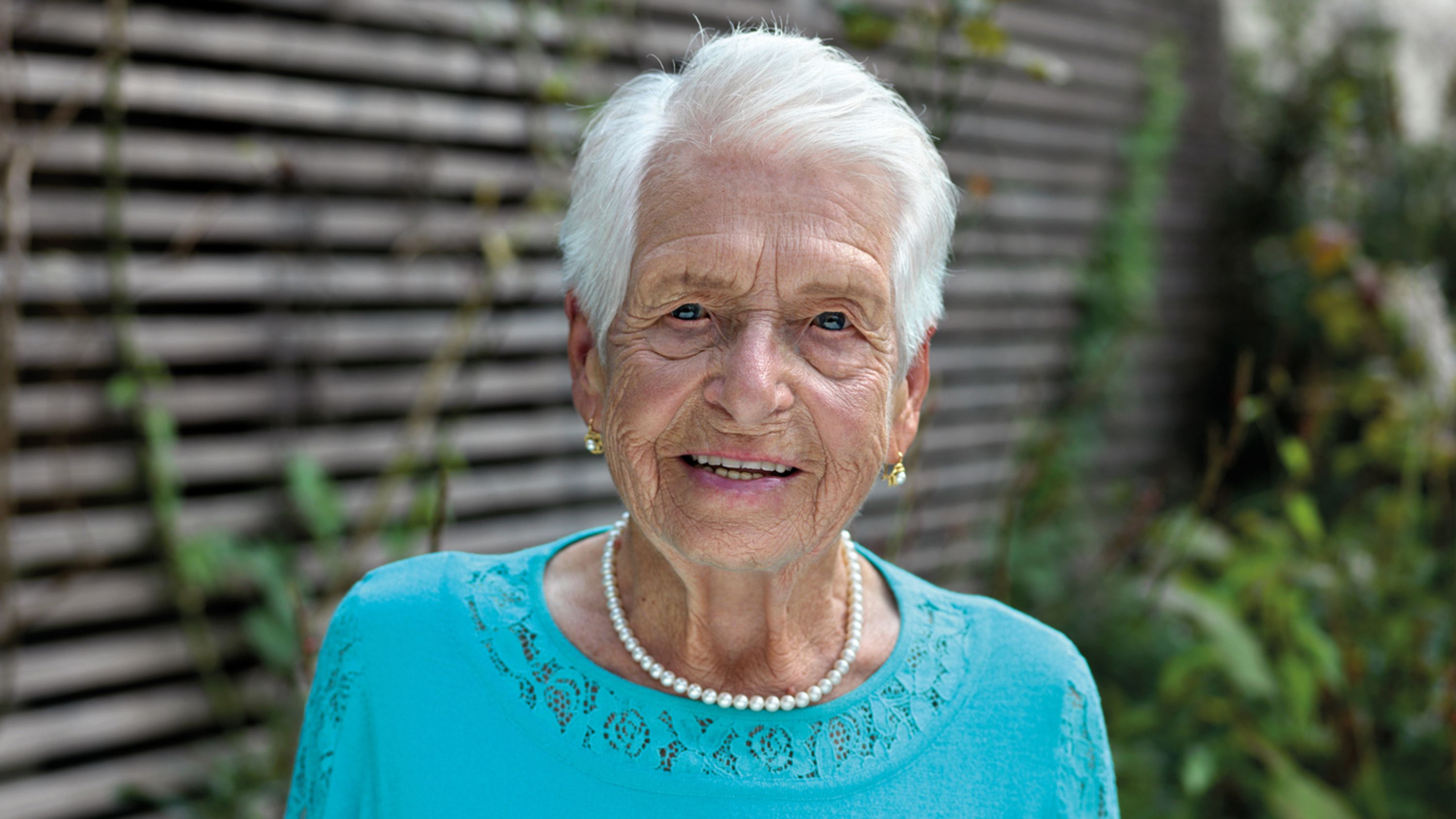 Une femme âgée portant un haut bleu et un collier de perles regarde la caméra en souriant.