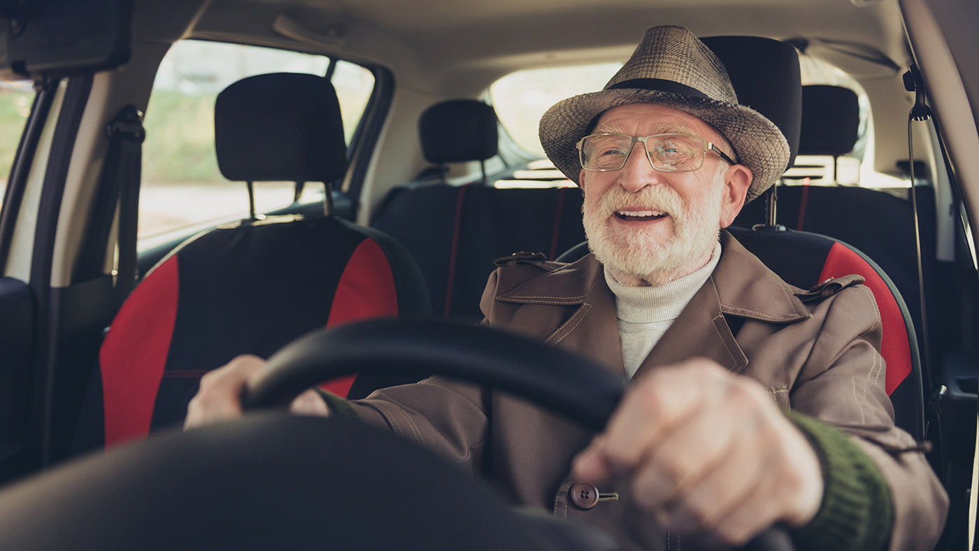 Un uomo anziano con occhiali, cappello e barba siede al volante e sorride.
