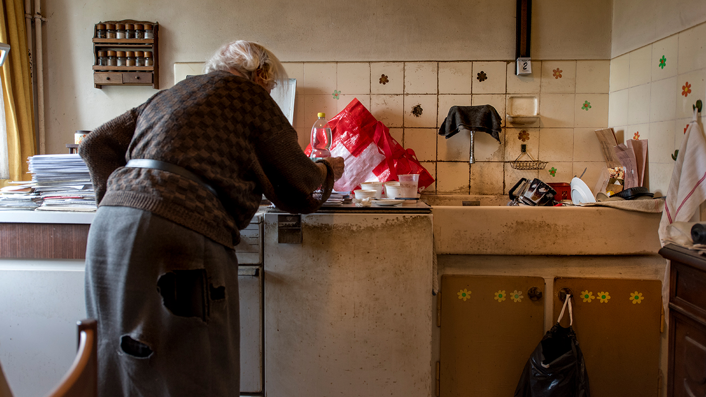 Une femme âgée vivant dans la pauvreté se tient debout, recroquevillée, dans sa cuisine.
